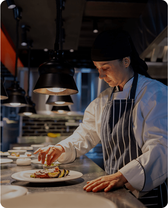 Chef plating dish under kitchen lights.